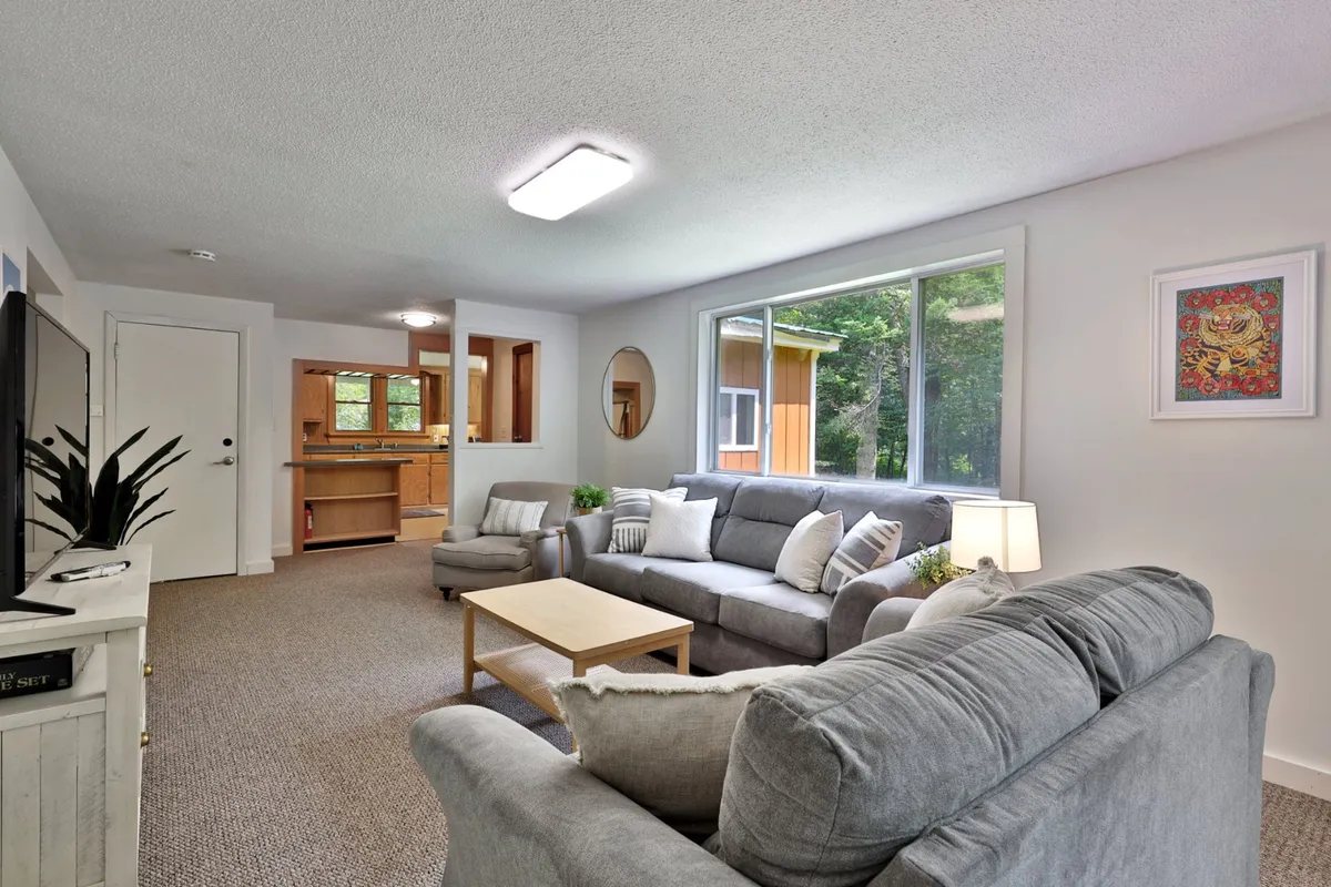 A bright living area with comfortable gray sofas, a wooden coffee table, and a large window overlooking greenery. The setting is cozy and welcoming, suggesting a place where guests can relax after outdoor adventures, regardless of the season.