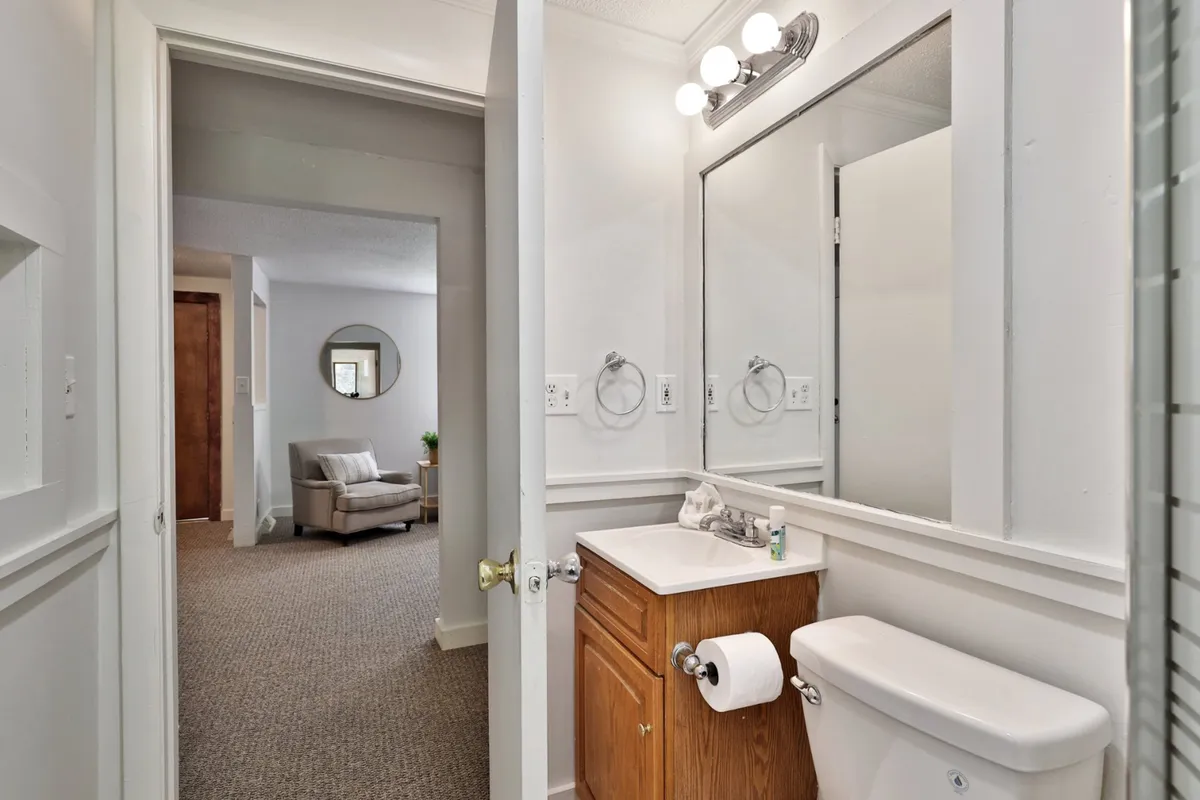 This image features a vanity with sink, a large mirror, a toilet, and a view through the doorway into a living area. The setting is neutral and inviting, showcasing a transitional space that highlights practical, everyday convenience and hints at the home's thoughtful design.
