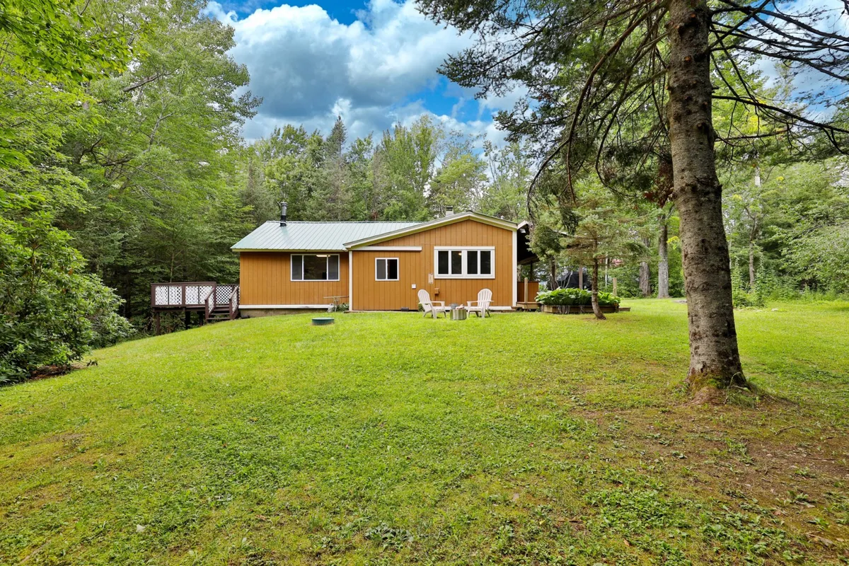 A yellow house with a large wooden deck, green lawn, and surrounding trees. The deck is furnished with outdoor seating, providing ample space for gatherings and barbecues. The peaceful scene is perfect for both daytime and evening activities, with open access to nature.