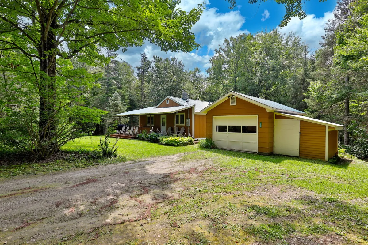This image features the yellow Mendon cottage nestled among trees, with a wide porch outfitted with comfortable chairs, inviting guests to relax after an active day. The house is surrounded by lush greenery, giving a sense of seclusion and a welcoming retreat from the slopes.