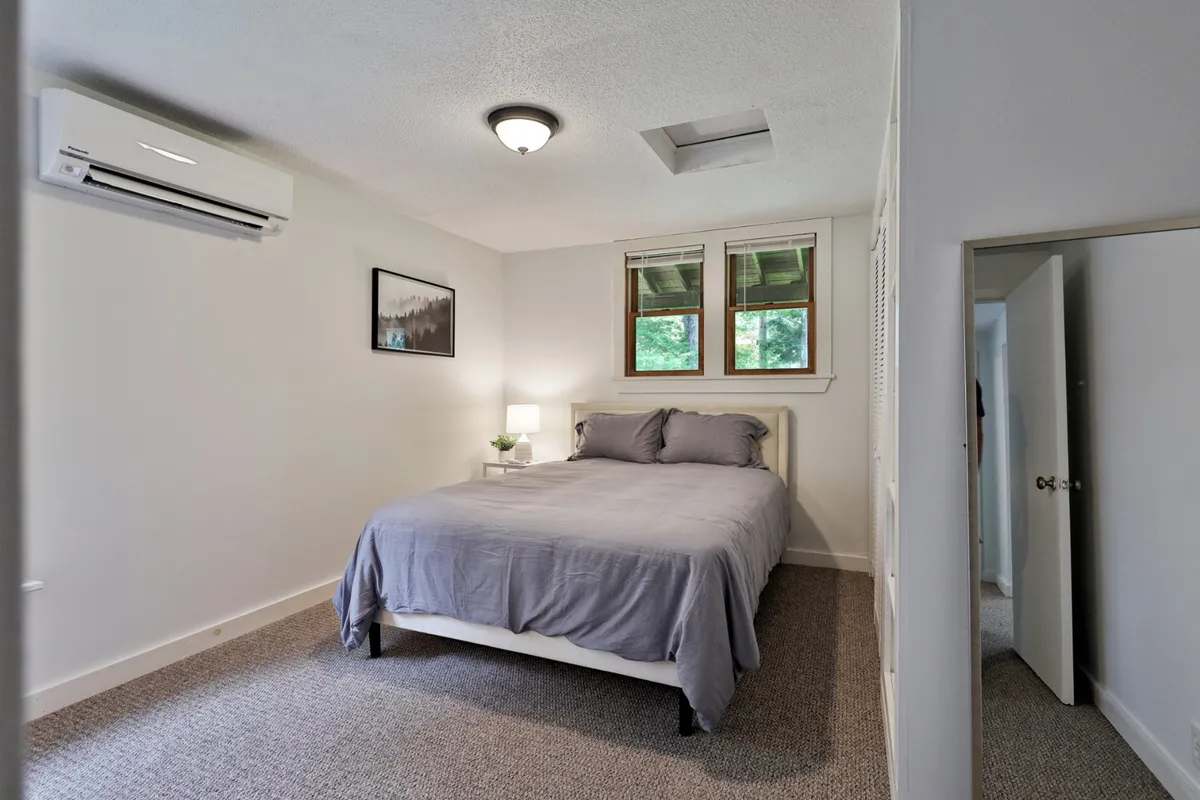 A serene bedroom featuring a plush bed with gray bedding, a table lamp paired with a small plant, and calming framed artwork on the wall. Natural light gently illuminates the peaceful atmosphere, underscoring the room's role as a restful retreat.