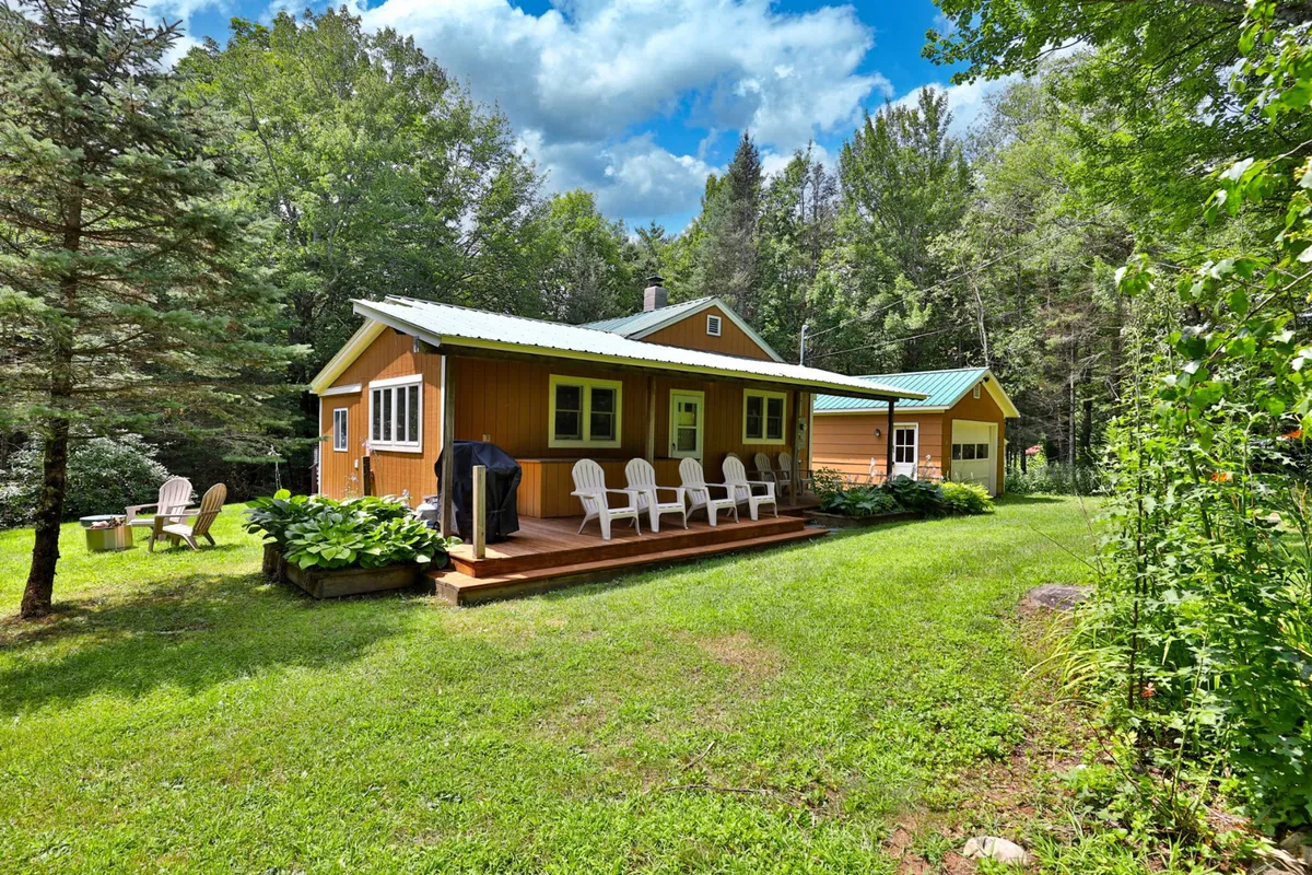 A spacious wooden deck bordered by green grass and surrounded by mature trees. White Adirondack chairs are set up, facing the tranquil landscape, creating inviting spots for relaxation or conversation. The setting captures the warm glow of sunset, emphasizing the peaceful, scenic atmosphere.