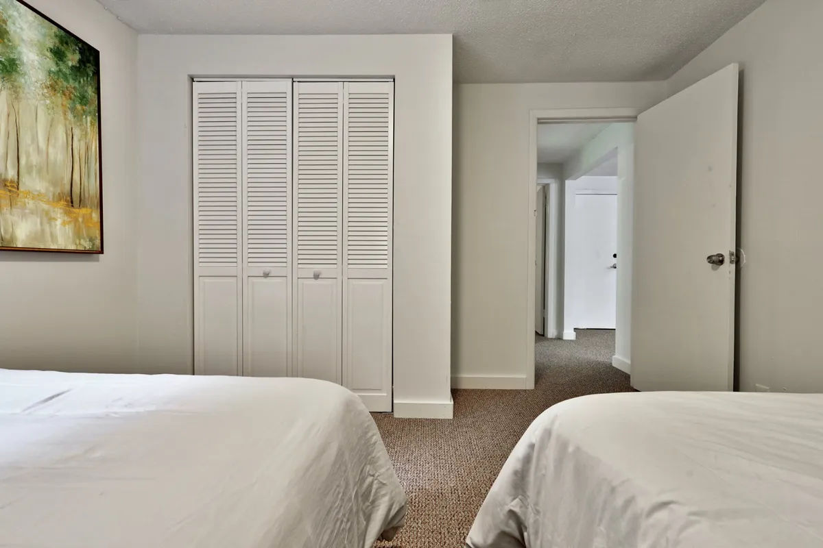 A tranquil second bedroom with two beds dressed in crisp white linens, a closet with louvered doors for storage, and a painting of trees on the wall. The room feels airy and calming, perfect for families or friends.