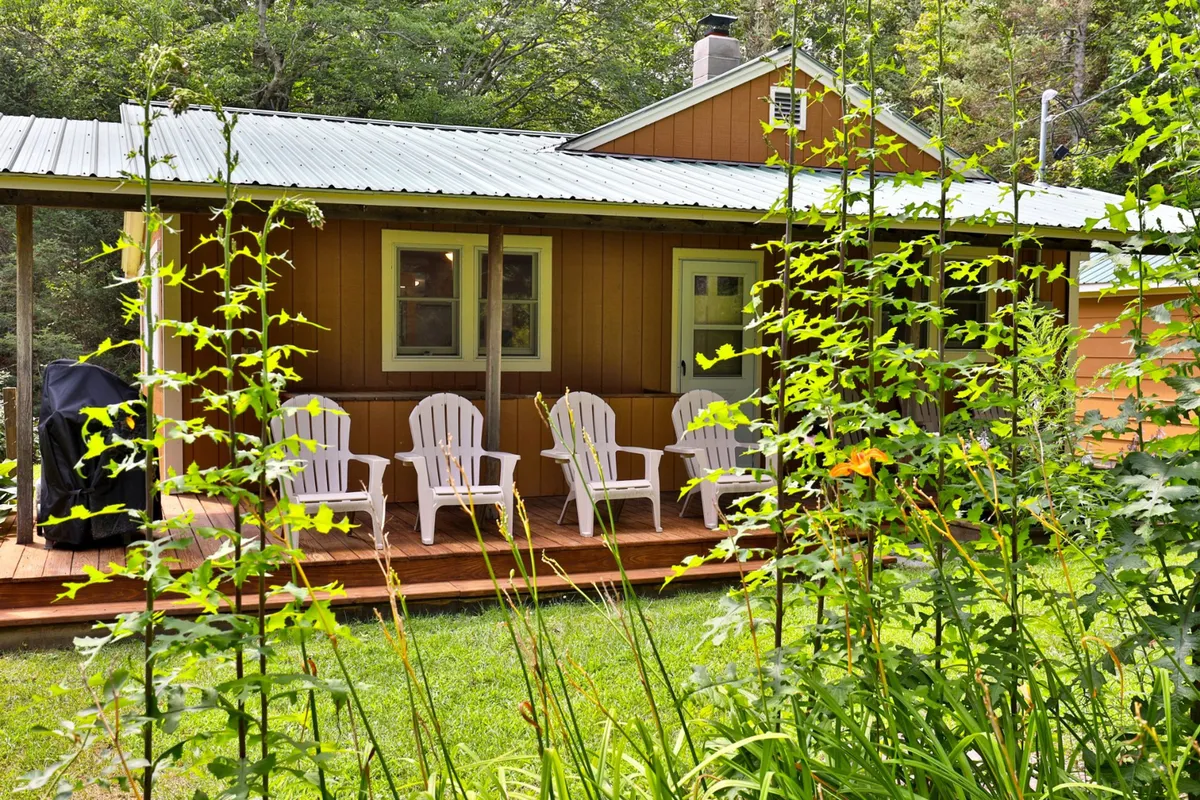White Adirondack chairs are set on a wooden deck adorned with yellow flowers and green foliage, with the brown exterior of the house in the background. The setup creates a cozy spot for relaxing, entertaining, and enjoying the natural surroundings throughout the day.