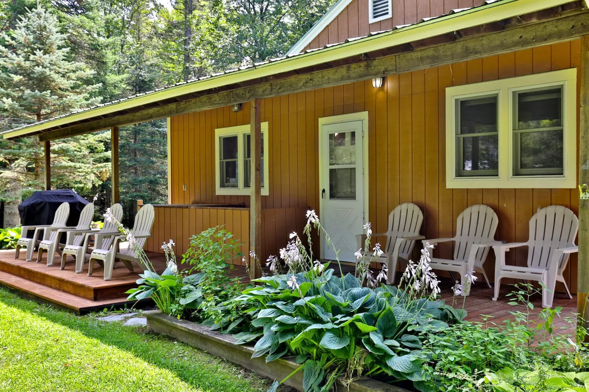 A charming exterior view of the wooden porch with Adirondack chairs, framed by lush greenery and vibrant flower beds. The inviting setup suggests a space ideal for unwinding, gathering with friends, and sharing stories under the stars.